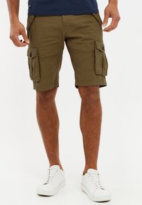 Mann trägt olivgrüne Cargo-Shorts mit Seitentaschen und weiße Sneaker vor einem weißen Hintergrund.