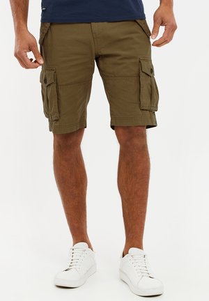 Homme portant un short cargo vert olive avec des poches latérales et des baskets blanches sur un fond blanc.