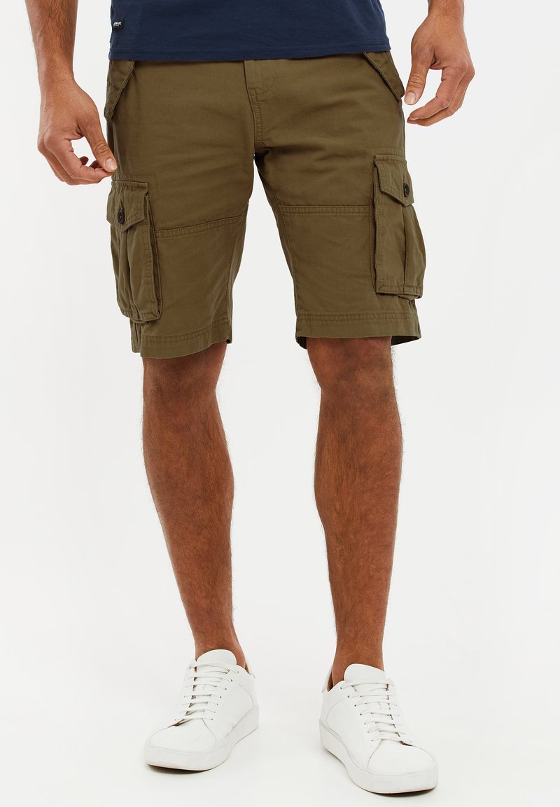 Mann trägt olivgrüne Cargo-Shorts mit Seitentaschen und weiße Sneaker vor einem weißen Hintergrund.