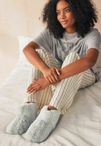 Faux fur hjemmesko i lys blå, med en blød tekstur og et hyggeligt design, parret med stribede lette bukser og en grå t-shirt.