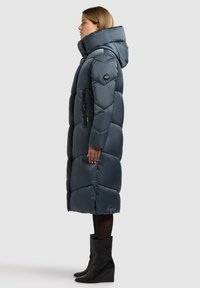 Pikk sinine üldmõõdus sulejope kapuutsiga ja külgmiste tõmblukuga taskutega. Omaduste hulgas on tikitud muster ja logodetail, sobitatud põlveni ulatuvate mustade kiilkontsaga saapadega.