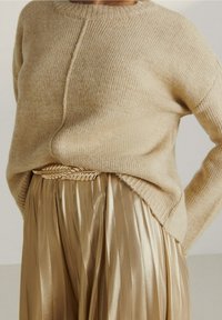 Beżowy sweter o prążkowanej fakturze i okrągłym dekolcie w połączeniu z metalicznie złotymi spodniami plisowanymi, z plecionym detalem na pasku.