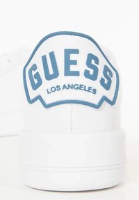 Sneaker bianco con grande etichetta blu "GUESS" con "LOS ANGELES" sottostante, su uno sfondo bianco liscio.