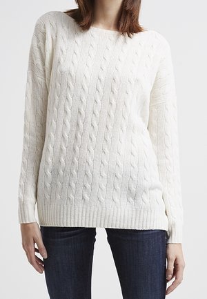 Woman wearing a white cable-knit sweater with long sleeves and dark blue jeans against a plain background.