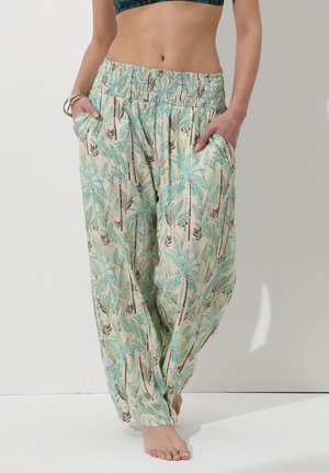 Donna che indossa pantaloni larghi a vita alta con stampa di foglie verdi e fiori, mani nelle tasche, in piedi a piedi nudi su uno sfondo neutro.