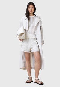 Witte trenchcoat over een witte textuur gebreide top en bijpassende shorts. Zilveren sandaal met bandjes. Gladde materialen, ontspannen pasvorm en verticale lijnen.