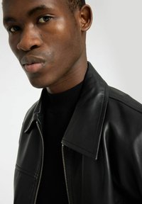 Schwarze Lederjacke mit spitzem Kragen, Reißverschluss und glatter Textur, getragen über einem schwarzen Stricktop. Das Gesicht des Models ist teilweise sichtbar.