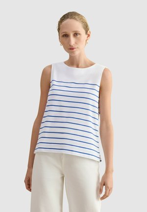 Mujer con cabello rubio que lleva una camiseta blanca sin mangas con rayas horizontales azules y pantalones blancos, de pie contra un fondo claro y sencillo.