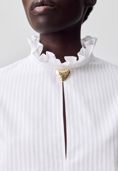 Primer plano de una persona que lleva una blusa blanca a rayas con un cuello rizado y un cierre en forma de corazón dorado en el escote.