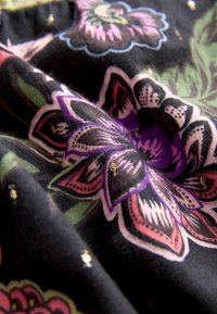 Tissu noir avec des motifs floraux vifs en rose, violet et vert, avec des accents dorés et une texture lisse.