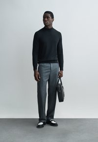 Uomo in piedi contro un muro semplice, indossa un dolcevita nero, pantaloni grigi, scarpe bianche e nere, tiene una valigetta nera nella mano destra.