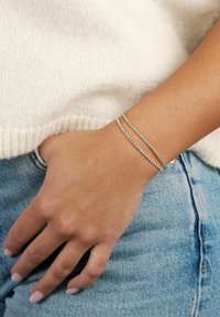 Armband i guldfärg med två strängar: en slät metall och en som har små klara kristaller, bärs på en hand som vilar på denim.