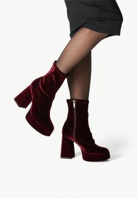Burgundowe aksamitne botki na kostkę z wysokim blokowym obcasem i bocznym zamkiem błyskawicznym, charakteryzujące się smukłym, zwężającym się fasonem i gładką teksturą.