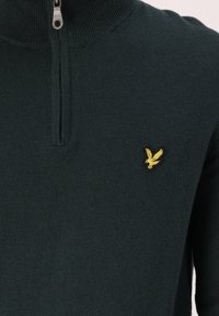Ciemnozielony sweter z zamkiem błyskawicznym przy kołnierzu, z małym, haftowanym żółtym logo ptaka po lewej stronie klatki piersiowej. Miękka tekstura.