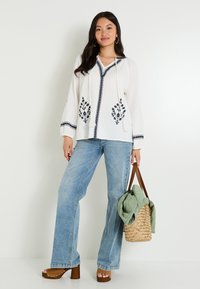 Blouse blanche à manches longues avec broderies florales bleu marine, décolleté en V, cordon de serrage, associée à un jean évasé bleu clair et des sandales à plateforme brunes. Sac tissé.