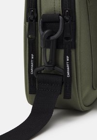 Zbliżenie narożnika zielonej torby z tkaniny z czarnymi suwakami oznaczonymi "Carhartt WIP" oraz czarnym plastykowym zaczepem mocującym czarny pasek.