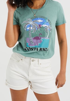 Kobieta w zielonym t-shircie z grafiką Kostaryki z palmami i flamingiem, w połączeniu z białymi szortami z podwiniętym dołem.