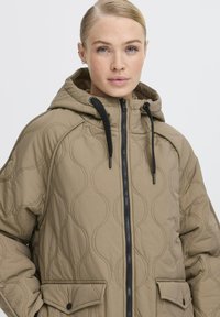 Beige gesteppte Kapuzenjacke mit Reißverschlussfront, schwarzen Beschlägen und Seitentaschen. Das Material weist strukturierte Wellenmuster auf.