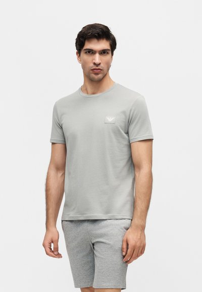 Άντρας που φοράει ανοιχτό γκρι κοντομάνικη μπλούζα με μικρό λογότυπο στο στήθος και γκρι σορτς ταιριαστά, σε απλό λευκό φόντο.