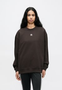 Καφέ φαρδιά φανέλα με ριμπ λαιμόκοψη και μανσέτες· διαθέτει λευκό λογότυπο Adidas στο στήθος· υφή υφάσματος λεία.