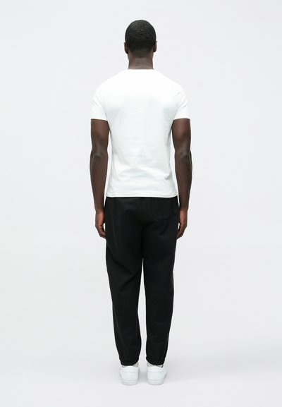 Mężczyzna stojący tyłem, ubrany w prostą białą koszulkę z krótkim rękawem, czarne spodnie z elastycznymi mankietami oraz białe trampki na białym tle.