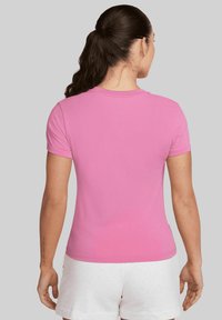 Donna con capelli scuri raccolti in un ponytail, che indossa una semplice maglietta rosa a maniche corte e pantaloni bianchi, in piedi con le spalle alla fotocamera.