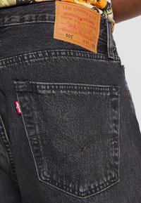 Vaqueros Levi's 501 negros que muestran el bolsillo trasero y el parche de cuero con la marca y la talla W31 L32 en una persona con una camisa estampada.