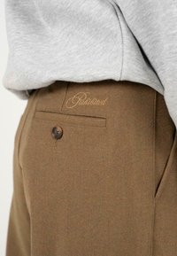 Pantalones marrones hechos de tejido texturizado, con un solo bolsillo trasero con un logo y cierre de botón. Parte de una blusa gris es visible.