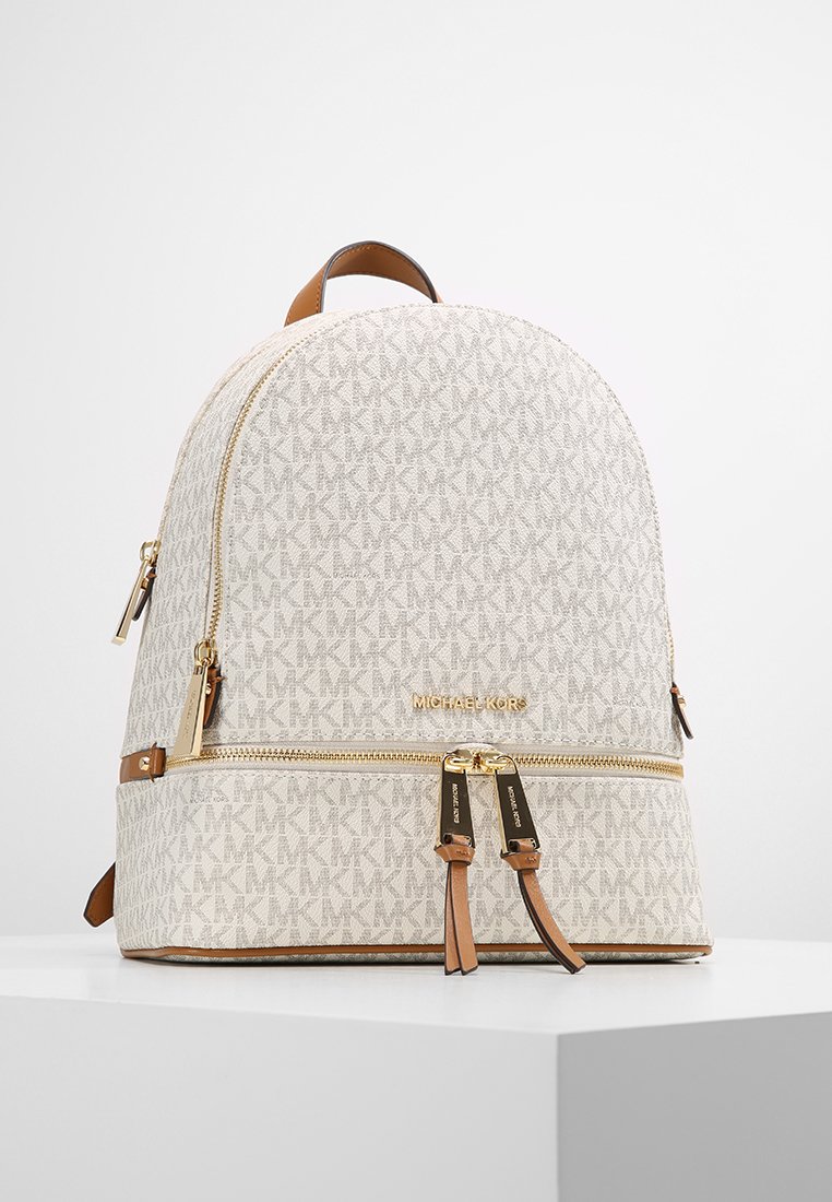 Witte canvas rugzak met patroon, gouden hardware, bruine accenten, een ronde bovenkant en dubbele ritsafsluitingen. Voorzien van een logo aan de voorkant.
