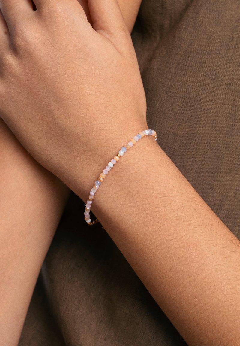 Pulsera delicada con pequeñas piedras preciosas multicolores en tonos pastel, montada en una cadena delgada, llevada en una muñeca desnuda sobre una tela texturizada.