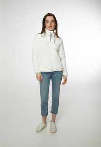 Giacca in pile bianca con colletto alto, zip frontale e due tasche laterali. Il modello indossa jeans azzurri chiari e sneaker bianche.