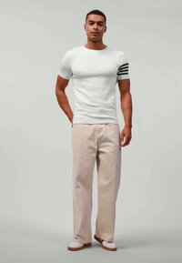 Maglietta bianca a righe con tre strisce nere sulla manica sinistra, abbinata a pantaloni larghi beige chiaro e calzature chiare. Design semplice, vestibilità casual.
