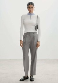 Witte langemouwen top met een paarse kraag en rits, gecombineerd met lichtgrijze geplooide broek en zwarte loafers. Kleine zwarte tas als accessoire.
