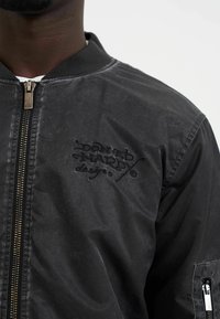 Black bomber jacket with a matte finish, ribbed collar, gold zipper, and embroidered "Donald Hardy designs" logo on the front.
