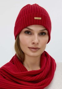 Red ribbed knit beanie with a leather logo tag, paired with a matching ribbed knit scarf, both featuring a soft texture.
