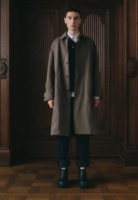 Brauner langer Mantel, weißes Hemd, schwarze Weste, marineblaue Hose und schwarze Gummistiefel mit roten Anhängern. Glattes Material, Knopfverschluss, entspannter Schnitt.