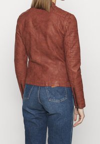 Kobieta ubrana w dopasowaną brązową kurtkę z zamszu z detalami zamka na rękawach oraz w spodnie dżinsowe o wysokim stanie w kolorze niebieskim, pokazana z tyłu.