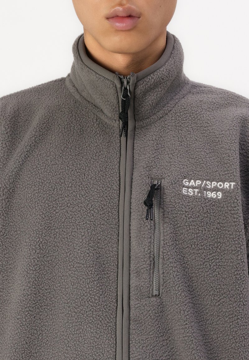 Grau Fleece-Jacke mit Reißverschluss vorne und hohem Kragen. Verfügt über eine Brusttasche und weißen Logo-Text "GAP/SPORT EST. 1969". Strukturierte Oberfläche.