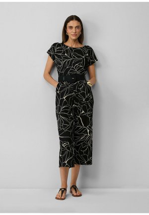 Femme debout contre un mur gris portant une combinaison noire avec des lignes florales blanches, une large ceinture, un bracelet et des boucles d'oreilles en or, ainsi que des sandales noires.