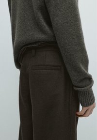 Ciemnoszary sweter z prążkowanymi mankietami, noszony na brązowych dopasowanych spodniach z pojedynczą kieszenią z tyłu i gładką fakturą.