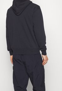 Man i en mörk marinblå huvtröja och matchande lösa byxor, ståendes vänd bort från kameran mot en enhetlig ljus bakgrund.