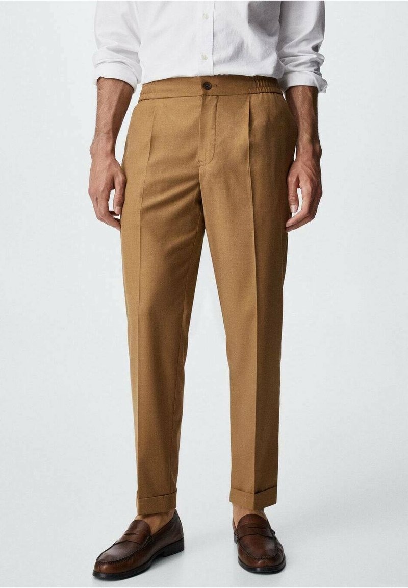 Braune maßgeschneiderte Hose mit glatter Textur, ausgestattet mit einem einzigen Knopfverschluss und vorderen Falten. Kombiniert mit dunklen Leder-Mokassins.
