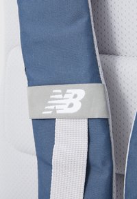 Sac à dos New Balance en tissu bleu avec des accents en maille blanche. Dispose d'une sangle grise avec un logo blanc et une surface texturée.