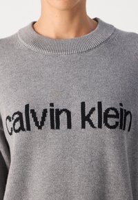 Hall lilla kudumüts ümmarguse kaelusega, mille ees on must "calvin klein" logo pikseliseeritud fontiga. Pehme tekstuur, lõdva disainiga.