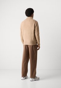 Beige strikket genser med høy krage, kombinert med løse brune bukser og grå joggesko. Teksturen er ribbestrikket og har et uformelt utseende.