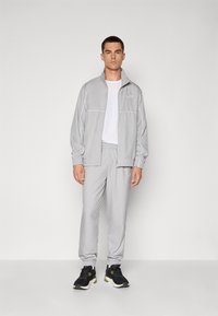 Lys grå træningsjakke og matchende bukser lavet af glat stof, med elastiske manchetter, lynlås-lommer og et logo på venstre bryst. Sorte sneakers.