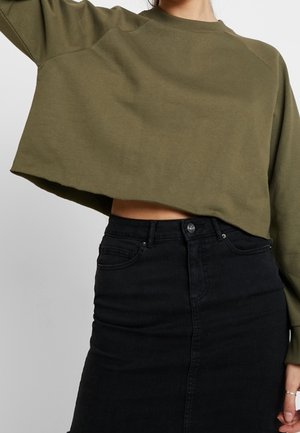 Oliwkowa krótka bluza zestawiona z czarną spódnicą z denimu z wysokim stanem odsłaniającą brzuch na neutralnym tle.