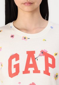 Krémová mikina s velkým červeným logem "GAP" a rozptýlenými květinovými potisky v růžové, žluté a modré barvě. Bavlněný materiál, kulatý výstřih.