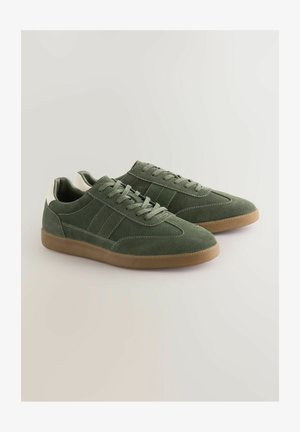 Žalios zomšinės sportbačiai su žemu profiliu, plokščiu gumuotu padu ir baltais akcentais ant kulno. Yra siuvinėjimo detalės ir raištelių uždarymas.