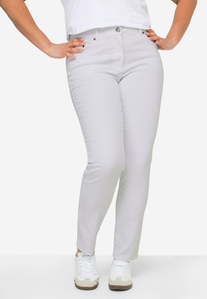 Frau trägt hellgraue, schlanke Hose, weiße Sneaker und ein weißes Oberteil, steht mit den Händen in der Hüfte vor einem einfarbigen Hintergrund.